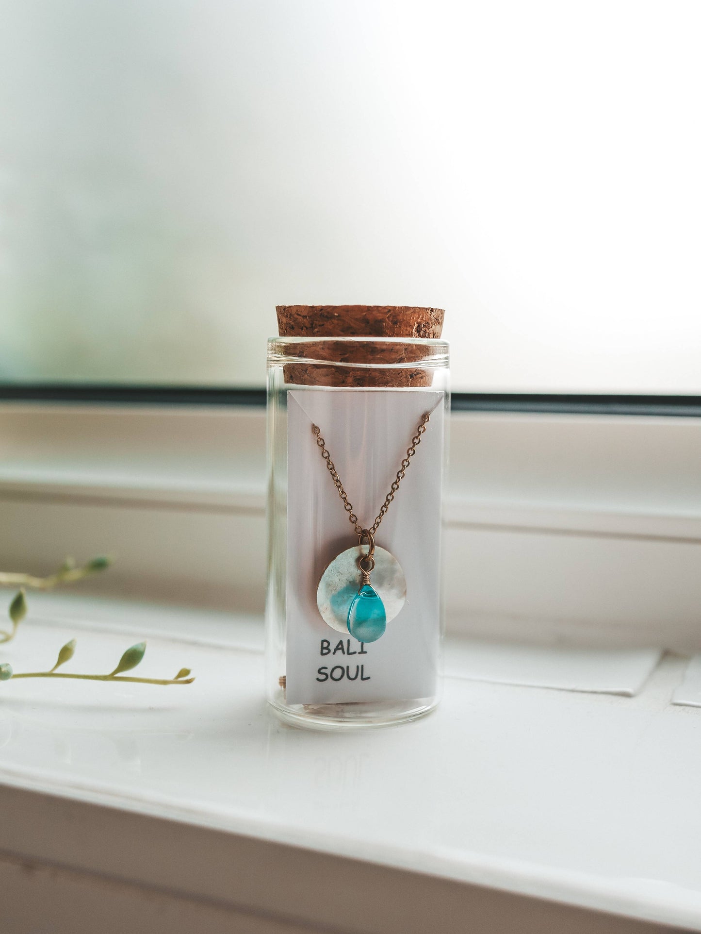 Necklace with a turquoise pendant in a glass jar labeled 'Bali Soul' on a windowsill.