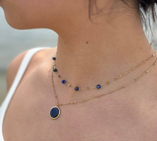 A close-up of a model's neck and upper chest wearing a double-layered gold necklace. The top layer is a delicate chain with small blue and gold beads, and the bottom layer features an oval gold pendant with a deep blue stone.