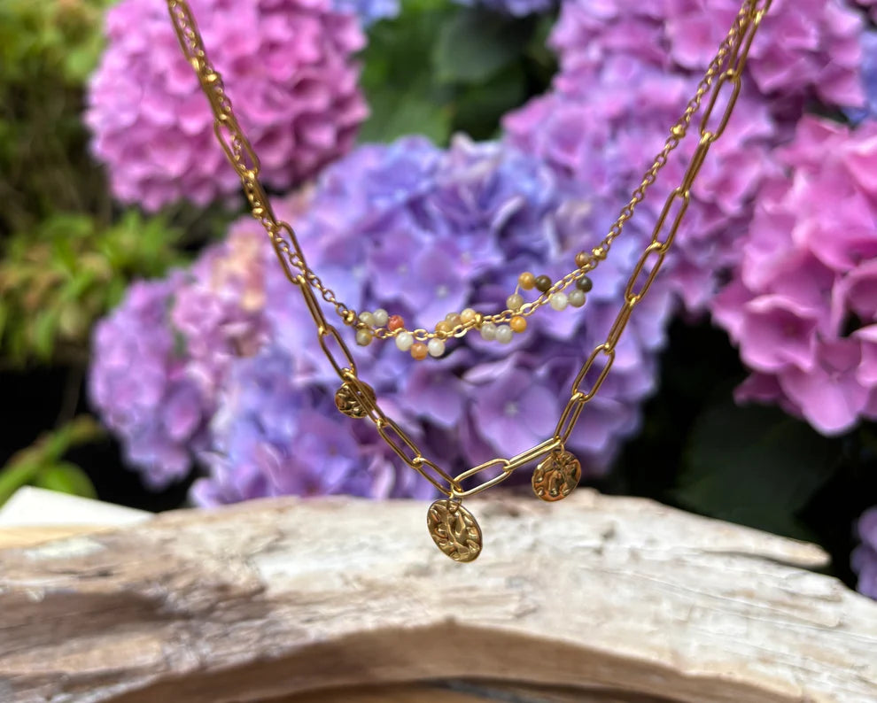 Gold necklace with small beads on a piece of driftwood in front of purple flowers