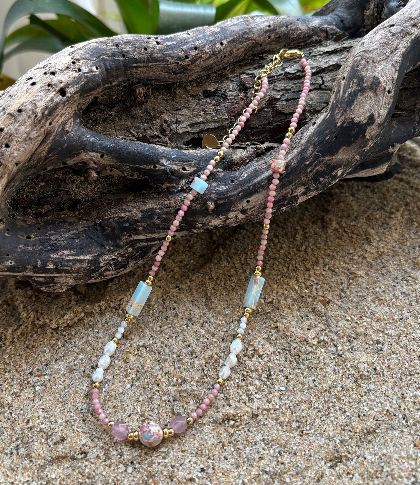 Beaded necklace with pink and blue beads on a sandy surface with driftwood in the background