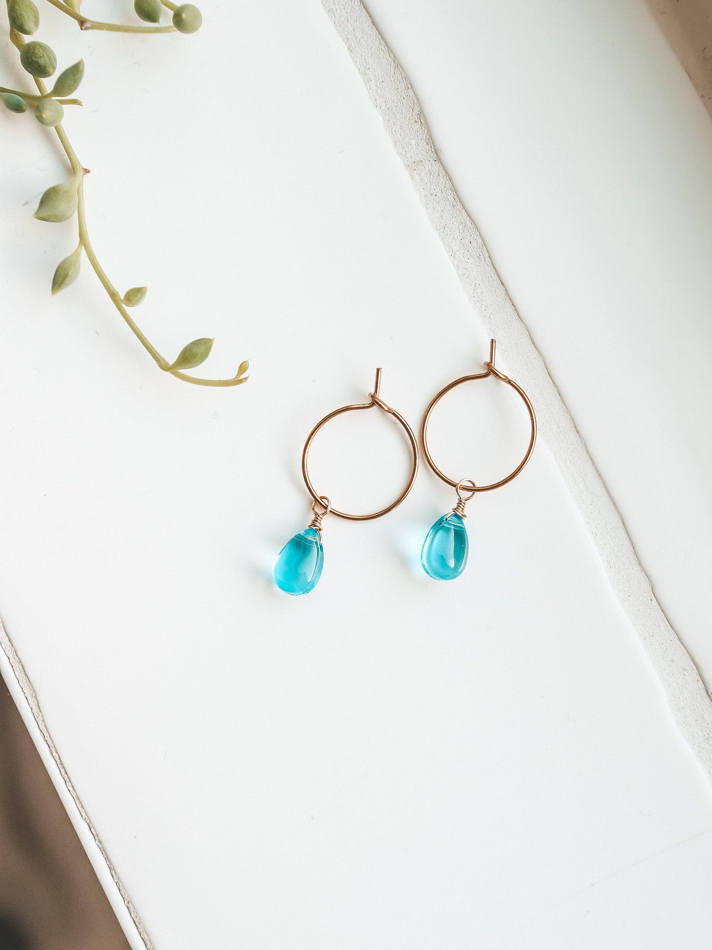 Gold hoop earrings with blue teardrop charms on a white surface with green leaves.