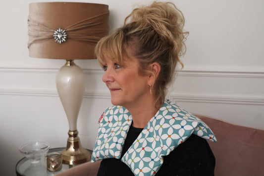 A woman with blonde hair in a messy bun wearing a patterned neck pillow, sitting on a pink sofa next to a lamp.
