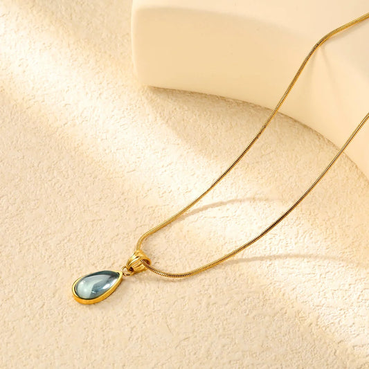 A gold chain necklace with a teardrop-shaped pendant featuring a light blue-gray stone in a gold bezel setting, displayed on a textured beige surface with soft shadows.