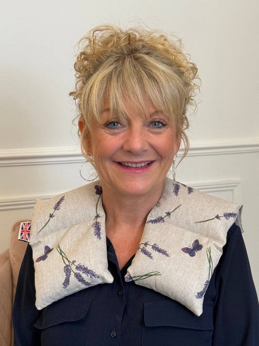 A smiling blonde woman wearing a padded lavender-patterned neck pillow with butterfly details over a navy blue collared shirt, set against a white paneled wall.