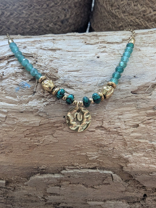 A beaded necklace with turquoise-blue beads, gold spacer beads, and a textured gold pendant, displayed on a rustic wooden surface.