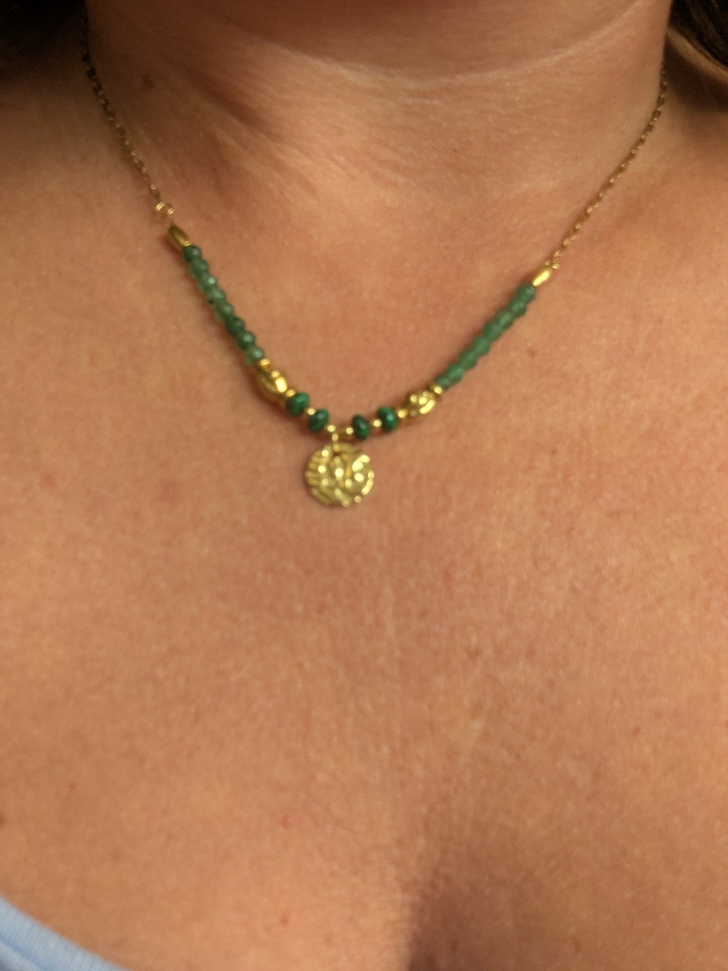 A close-up of a delicate necklace worn on a person's neck, featuring green faceted beads, small gold spacer beads, and a textured gold circular pendant.