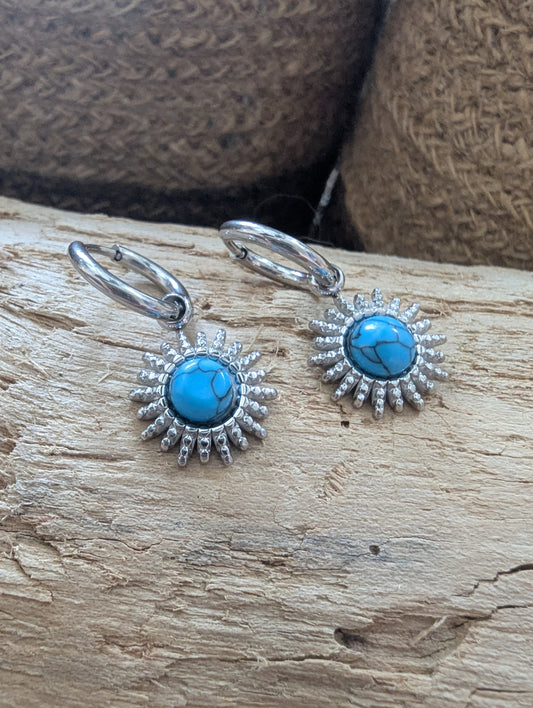 A pair of sterling silver drop earrings with round turquoise cabochons and sunburst detailing, displayed on a piece of natural wood with a woven brown background.