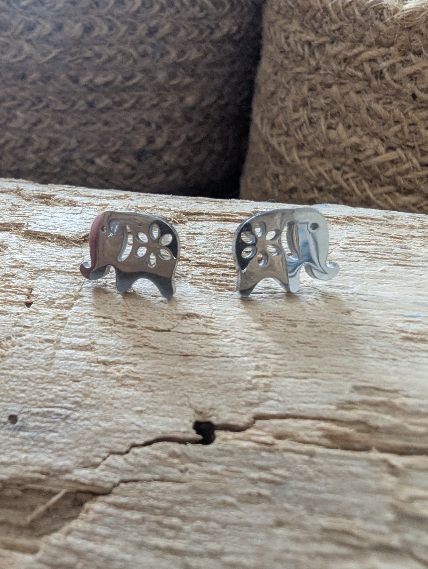 A pair of silver elephant-shaped stud earrings with cutout details, resting on a textured light wood surface with a woven brown background.