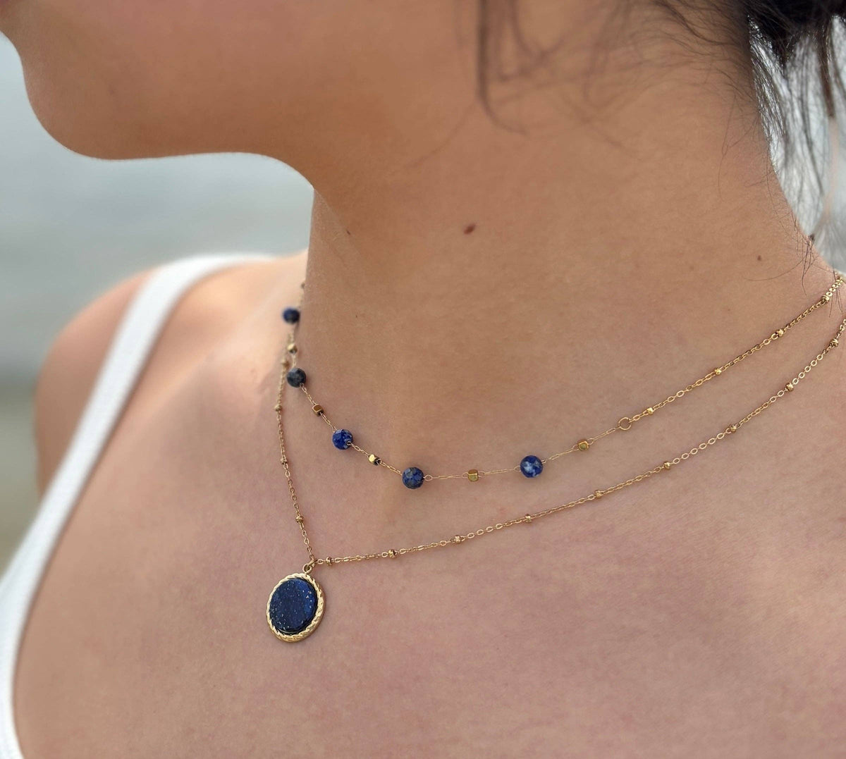 A close-up of a model's neck and upper chest wearing a double-layered gold necklace. The top layer is a delicate chain with small blue and gold beads, and the bottom layer features an oval gold pendant with a deep blue stone.