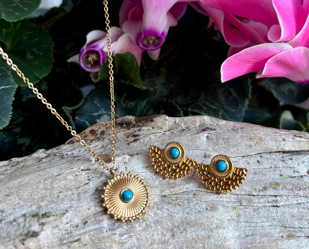Gold necklace and earrings with turquoise stones on a wooden surface with pink flowers in the background