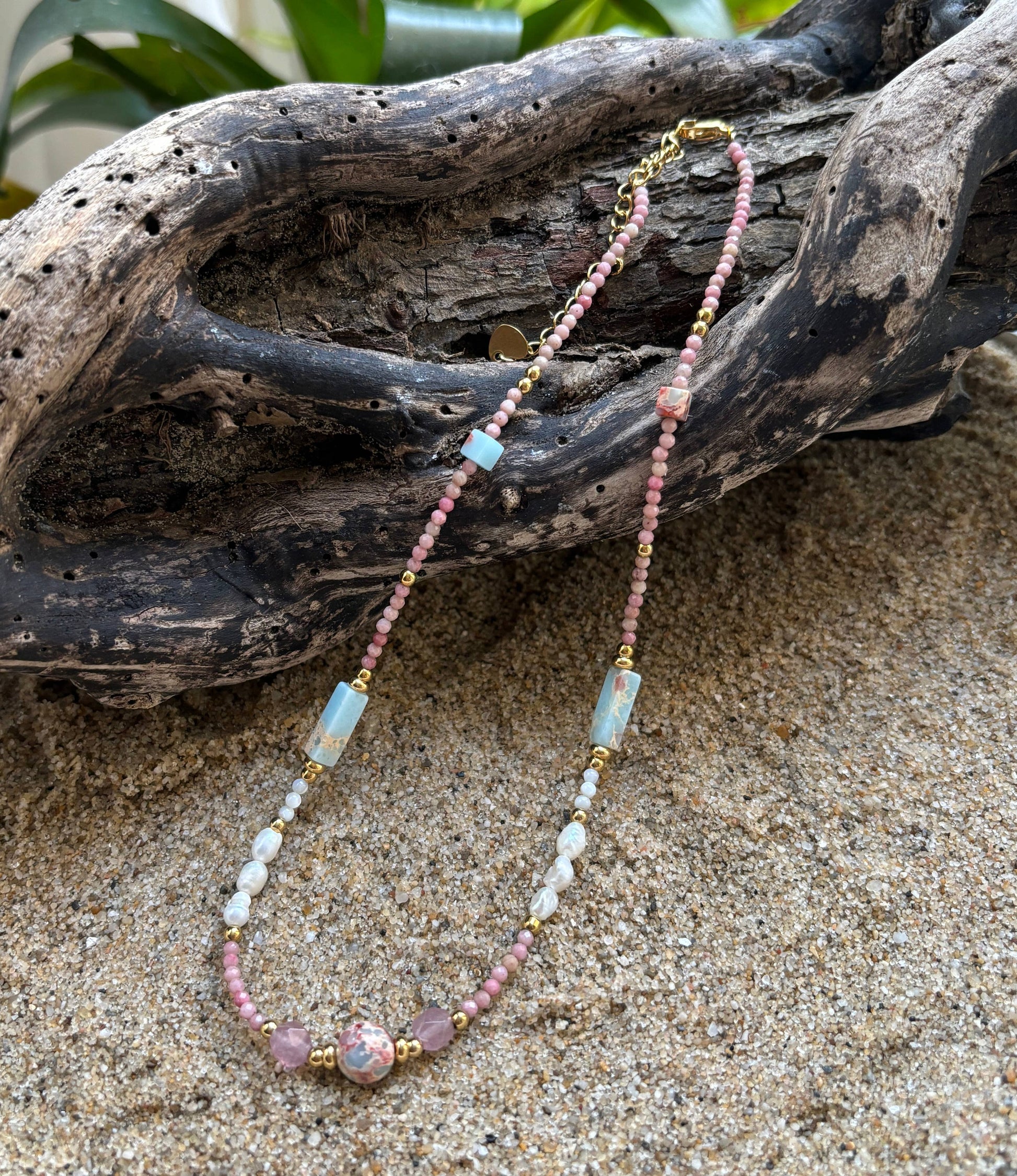Beaded necklace with pink and blue beads on a sandy surface with driftwood in the background