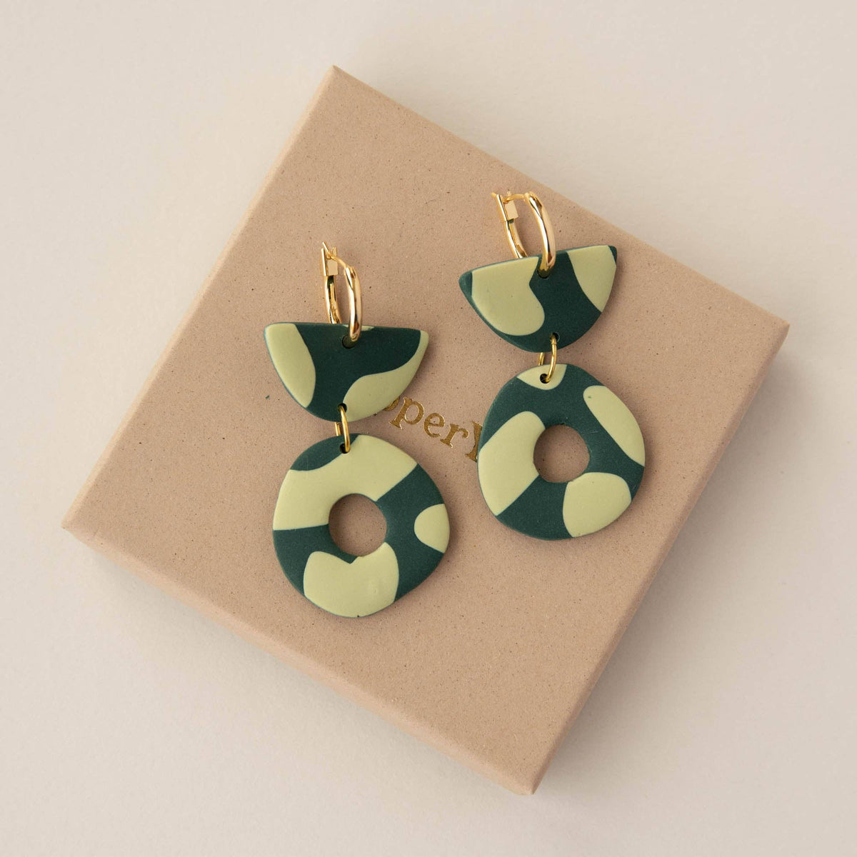 Green and beige patterned earrings on a brown box with 'per' branding.