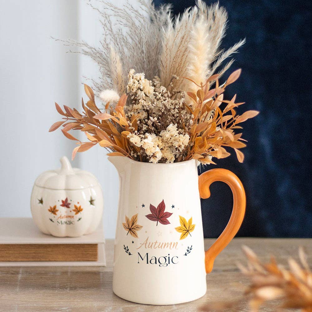 White pitcher with orange handle and 'Autumn Magic' text, filled with dried flowers on a wooden surface.