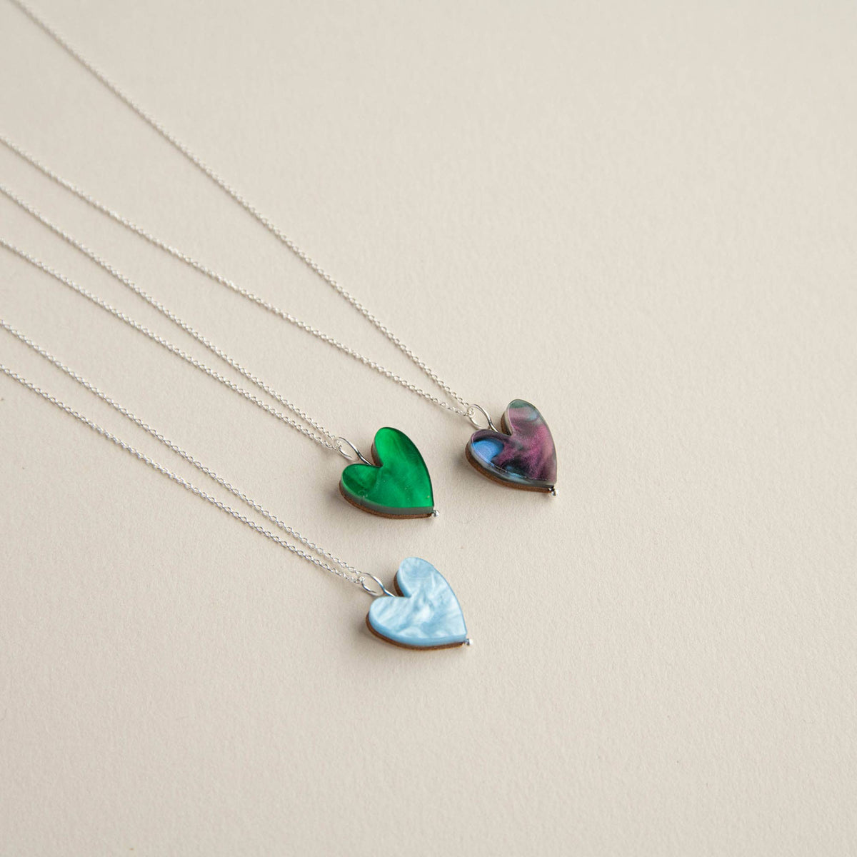 Three heart-shaped pendant necklaces on silver chains, displayed diagonally on a light beige background. The necklaces feature iridescent heart pendants in green, light blue, and a purple-blue marbled pattern.