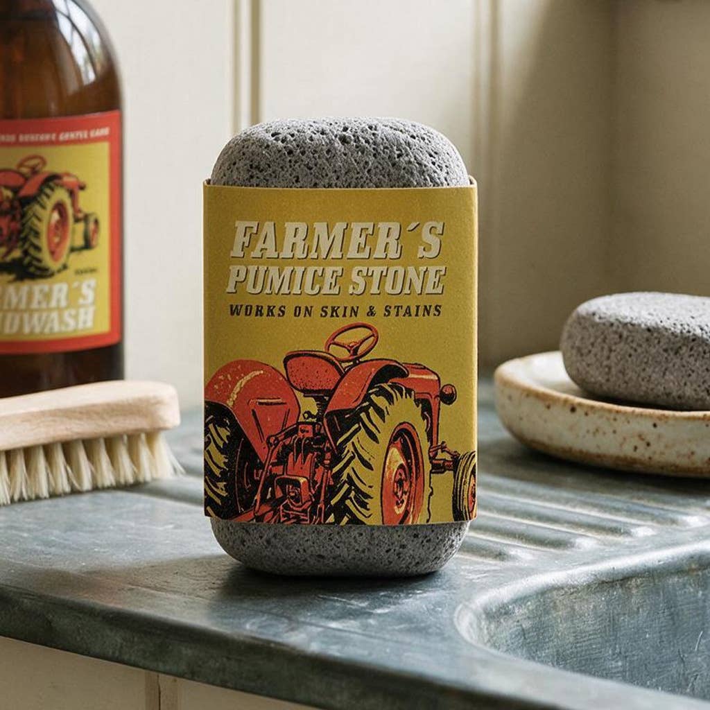 A pumice stone soap bar wrapped in a yellow paper sleeve featuring a red tractor illustration and the text 'FARMER'S PUMICE STONE' sits on a metal sink counter. A wooden brush, a brown bottle, and another soap bar are visible in the background.