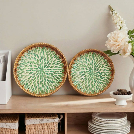 Two round woven decorative trays with green and cream radial patterns and natural wicker edges, displayed on a wooden shelf in a styled interior setting.