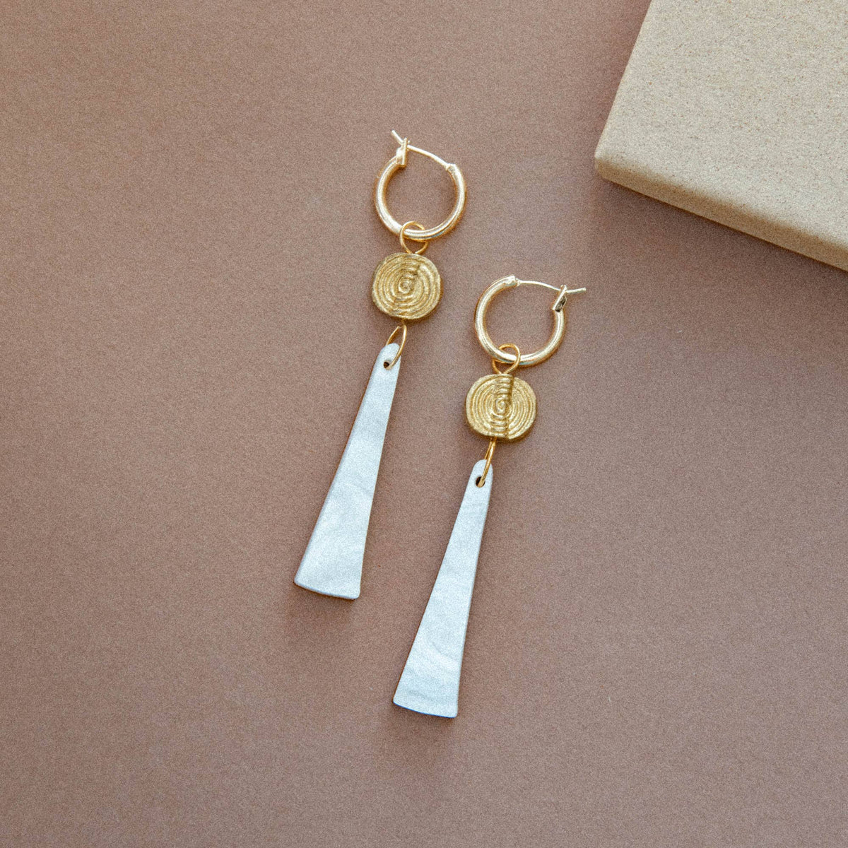 A pair of gold hoop drop earrings with textured circular discs and elongated white teardrop pendants, displayed on a warm brown background.