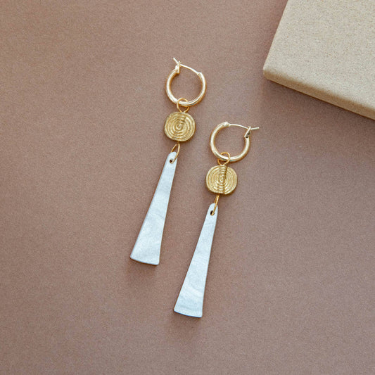 A pair of gold hoop drop earrings with textured circular discs and elongated white teardrop pendants, displayed on a warm brown background.