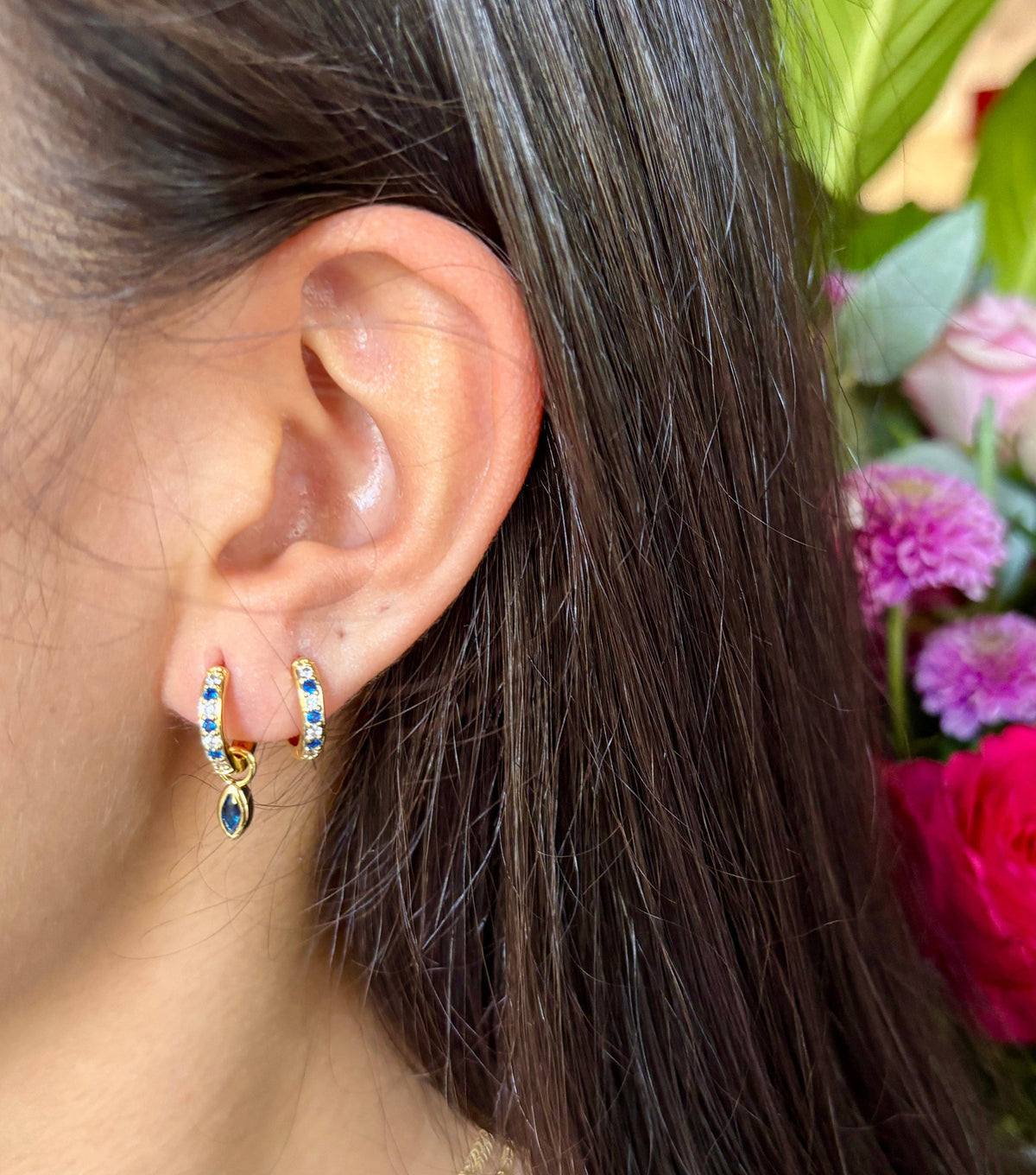 Close-up of an ear wearing a small hoop earring with colorful stones, surrounded by flowers.