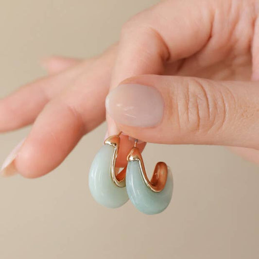 Hand holding a pair of light blue hoop earrings with gold accents against a beige background