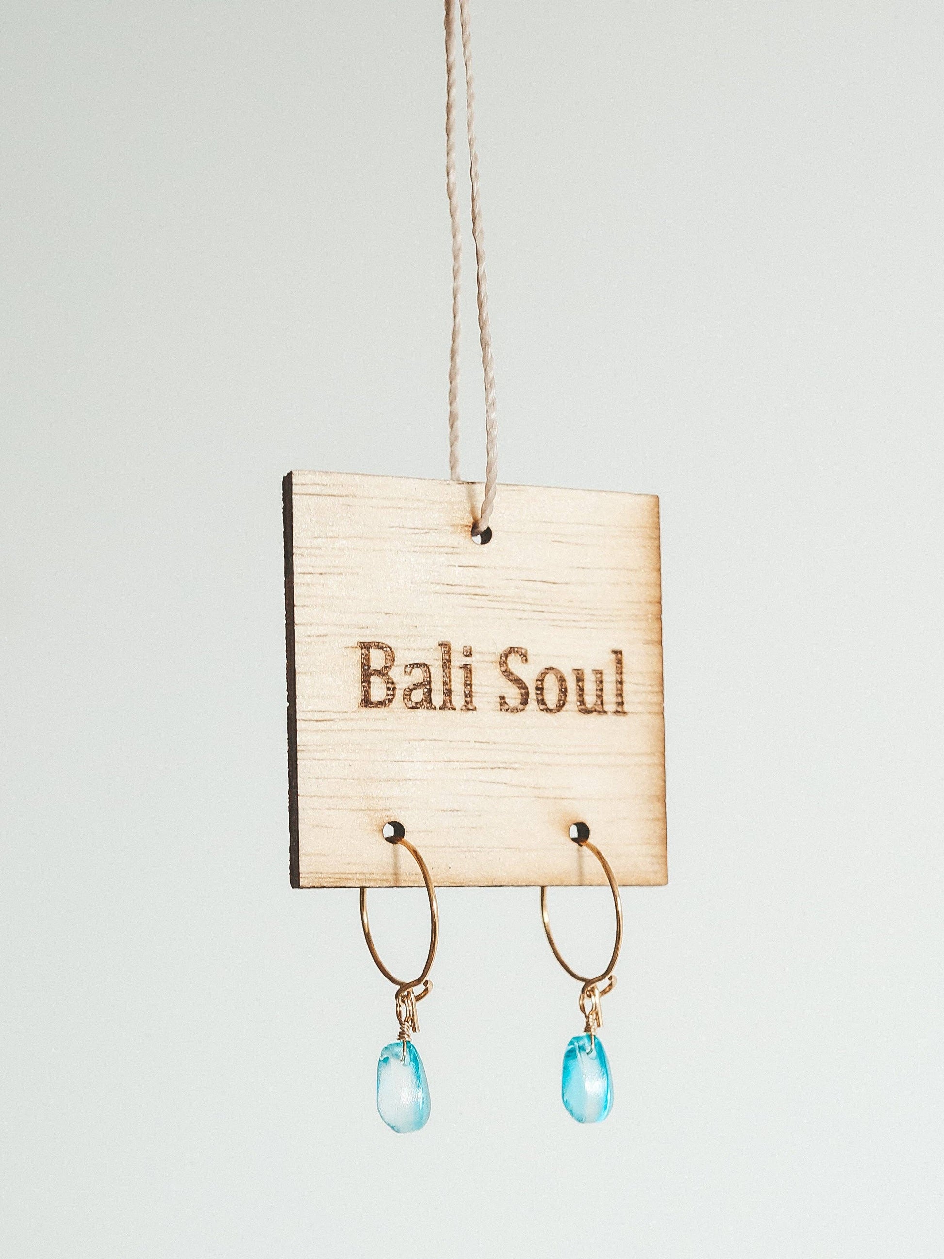 Earrings with blue drops hanging on a 'Bali Soul' sign against a light gray background