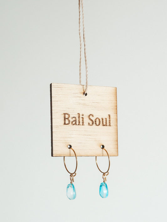 Earrings with blue drops hanging on a 'Bali Soul' sign against a light gray background