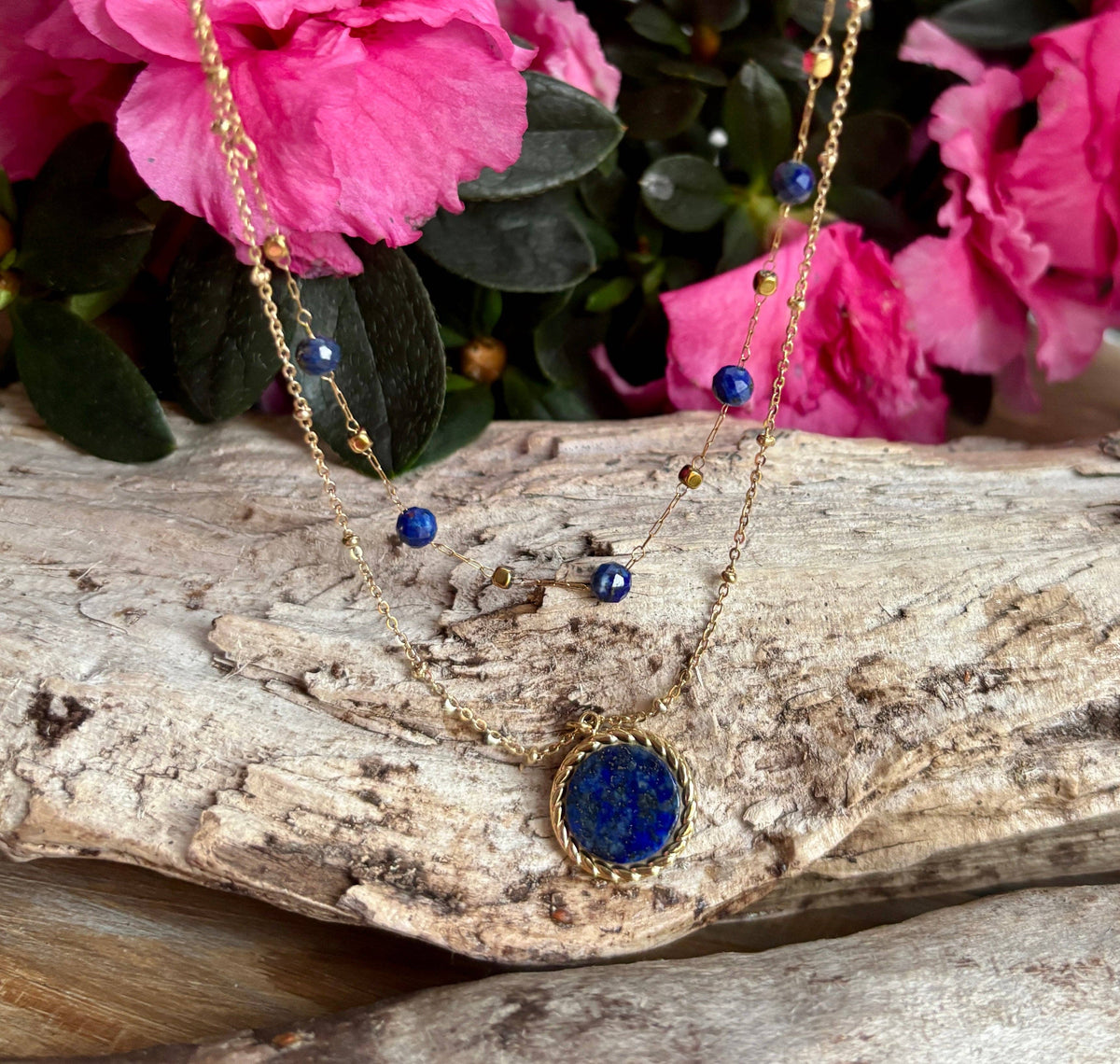 A gold-toned double-strand necklace with a round blue lapis lazuli pendant, displayed on a piece of driftwood with pink flowers in the background.