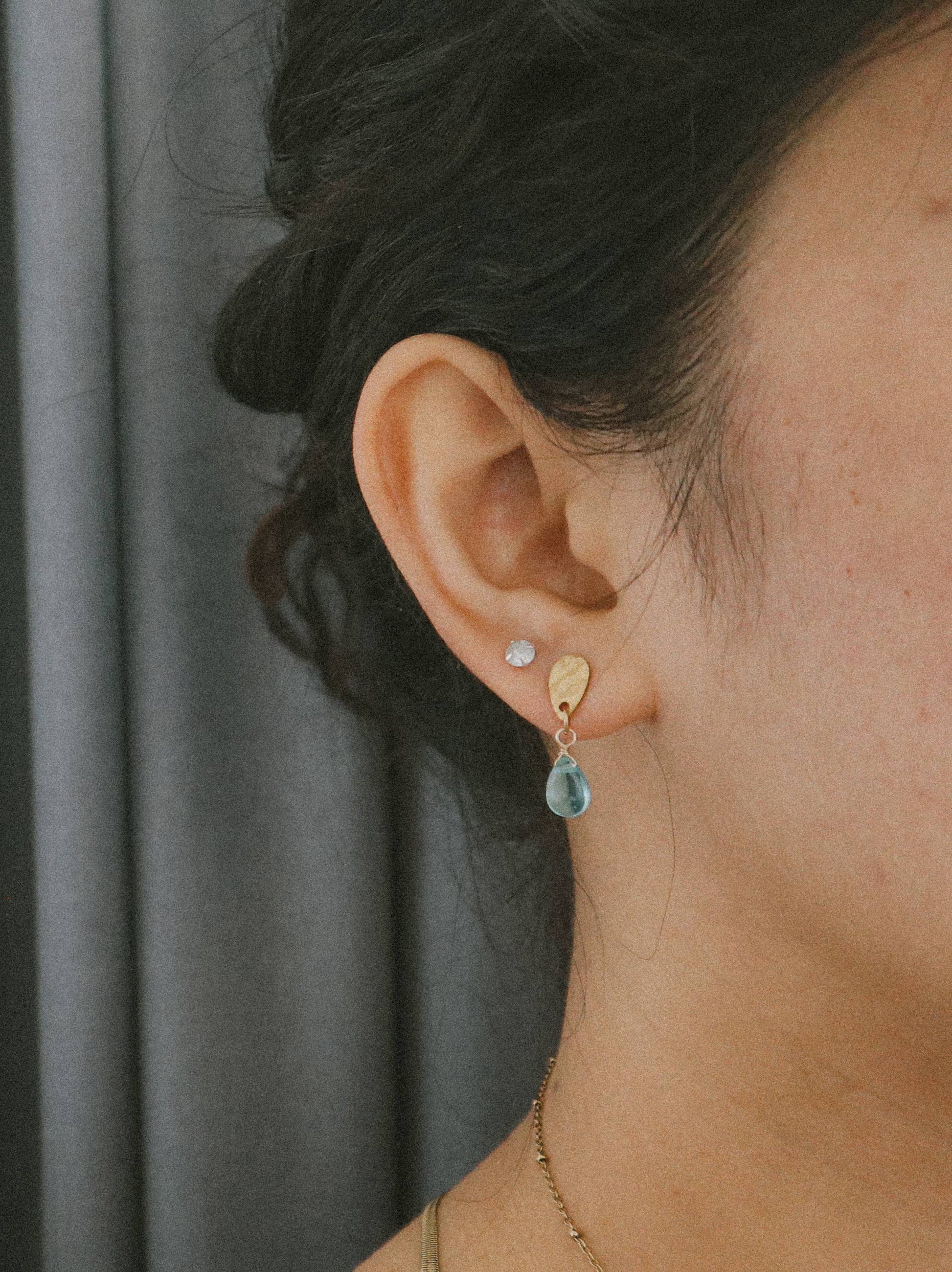 Close-up of an ear wearing a gold earring with a blue gemstone against a neutral background