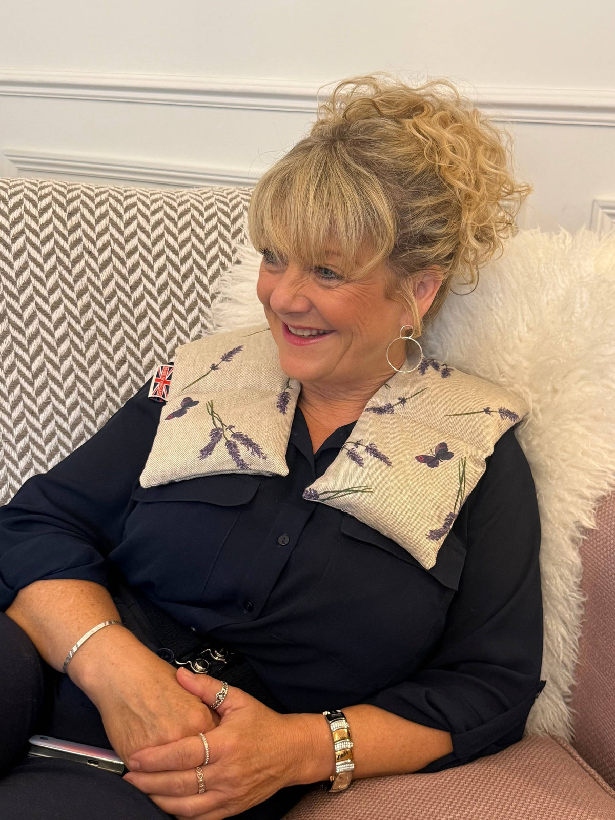 A smiling blonde woman sits on a patterned sofa wearing a navy blouse and a cream fabric neck pillow with purple floral and butterfly prints.