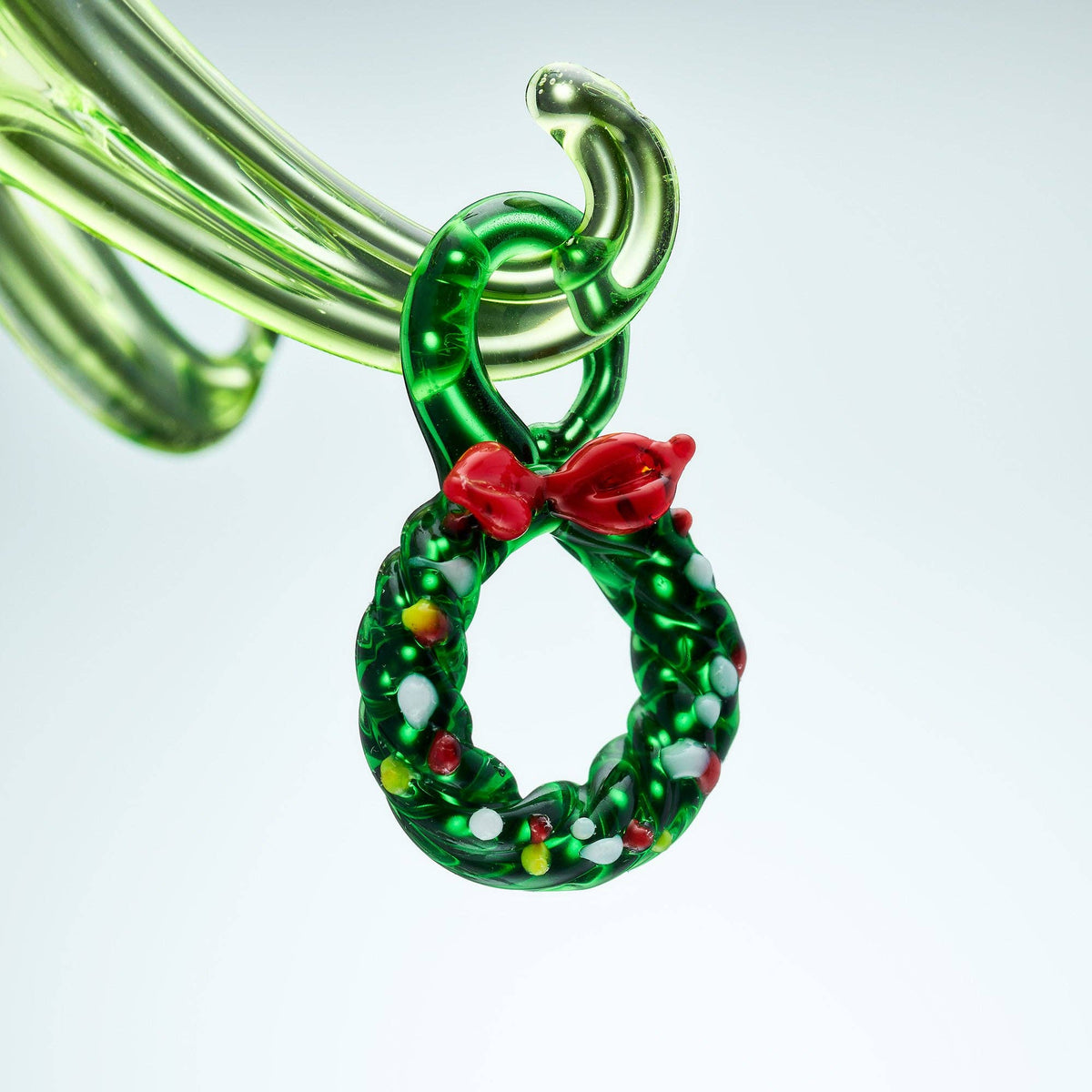 Decorative green glass ornament shaped like a wreath with a red bow on a light gray background