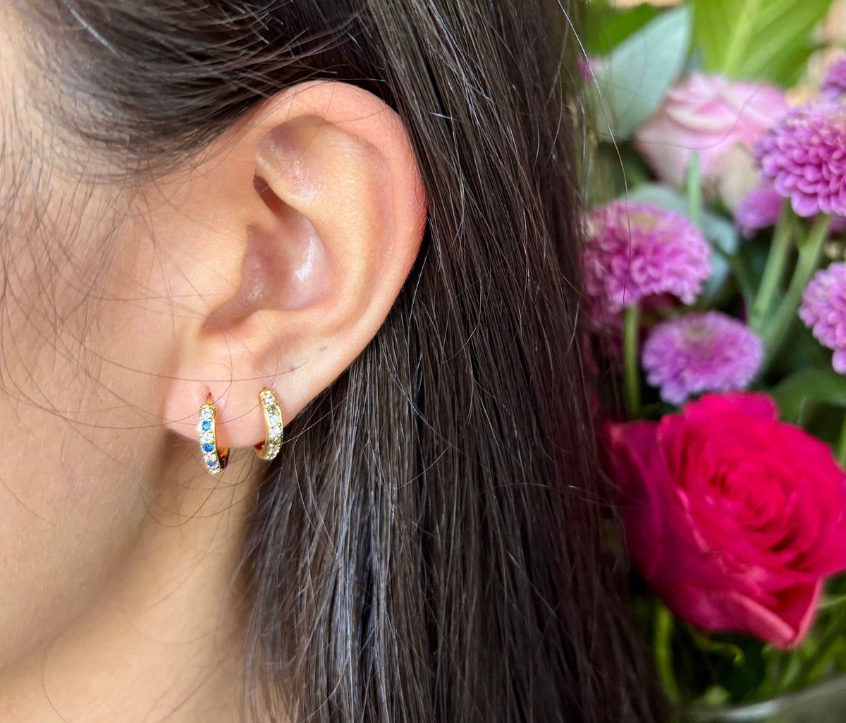 Close-up of an ear wearing gold hoop earrings with a bouquet of flowers in the background