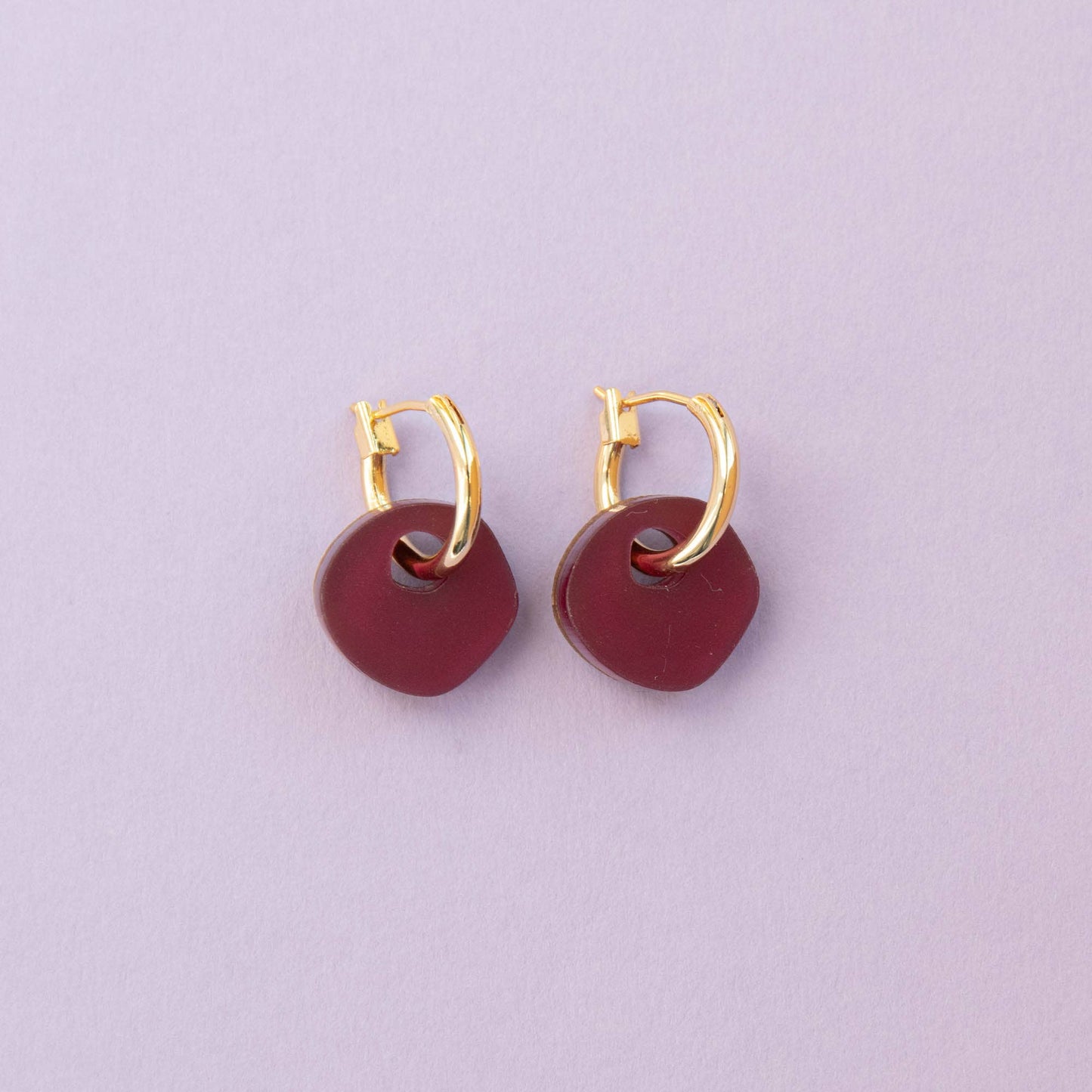 Gold hoop earrings with burgundy circular charms on a light purple background