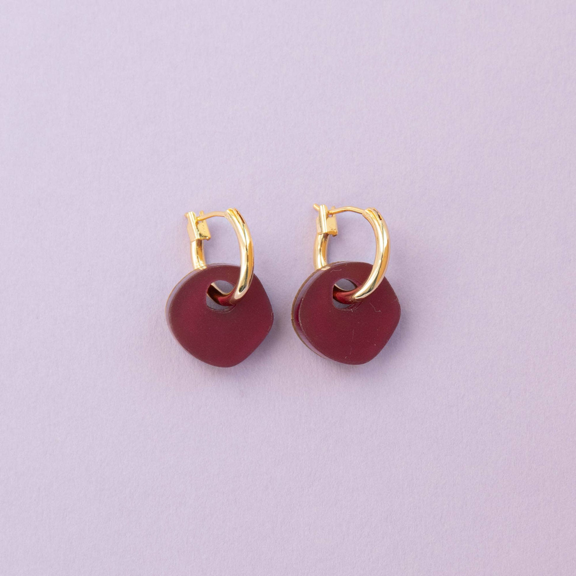 Gold hoop earrings with burgundy circular charms on a light purple background