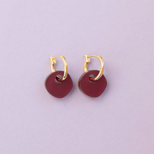Gold hoop earrings with burgundy circular charms on a light purple background