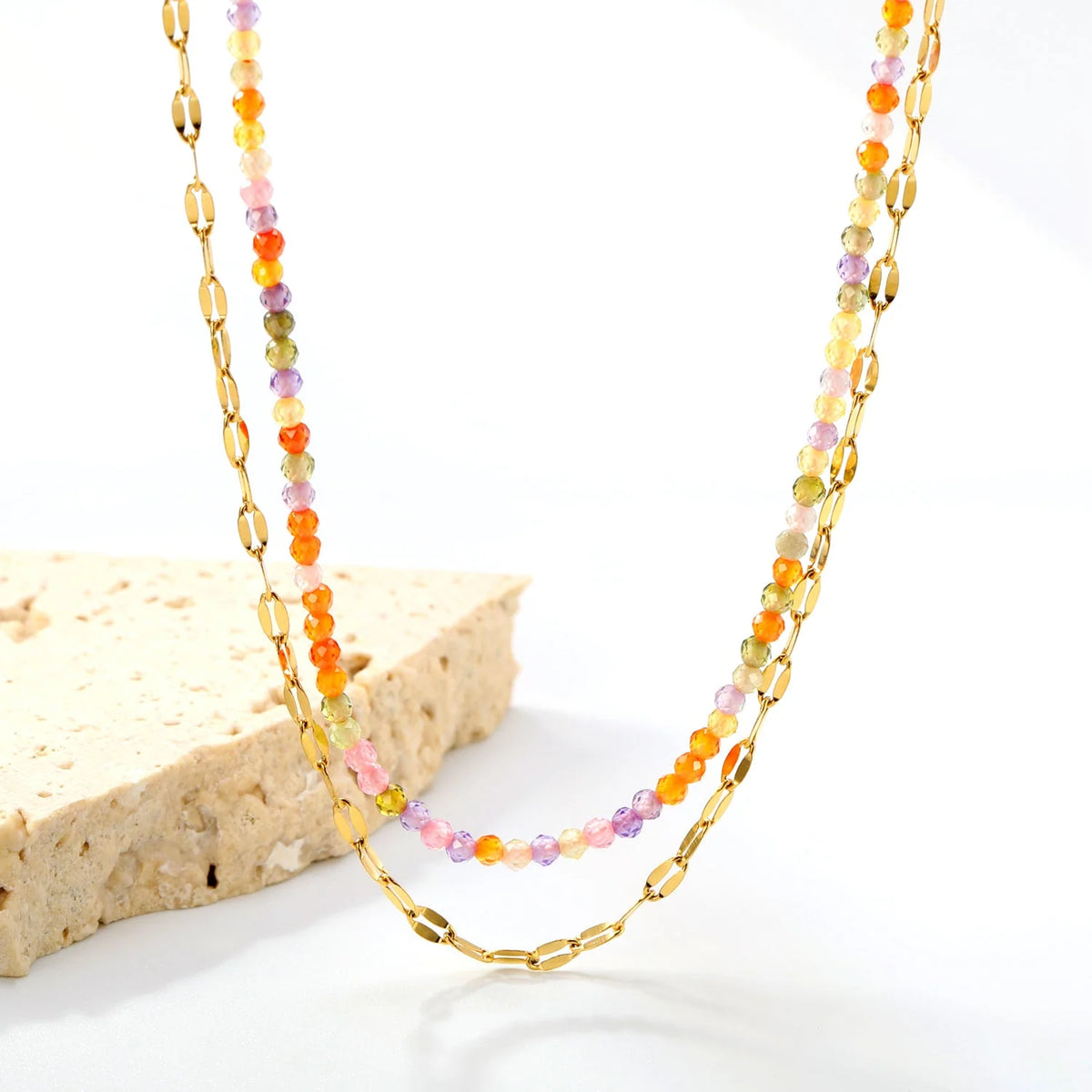 A double-strand gold chain necklace, one with elongated oval links and the other with small multicolored faceted beads, displayed on a white surface next to a light tan stone block.