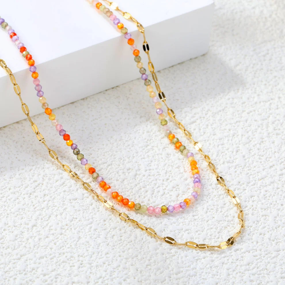 A double-strand necklace featuring a gold chain layered with a strand of small, faceted beads in pastel orange, pink, purple, and green, displayed on a textured white surface next to a white block.