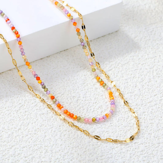 A double-strand necklace featuring a gold chain layered with a strand of small, faceted beads in pastel orange, pink, purple, and green, displayed on a textured white surface next to a white block.