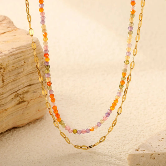 A double-strand necklace featuring a gold chain layered with small, colorful faceted beads in orange, purple, green, and pink, displayed on a textured beige surface next to a piece of driftwood.