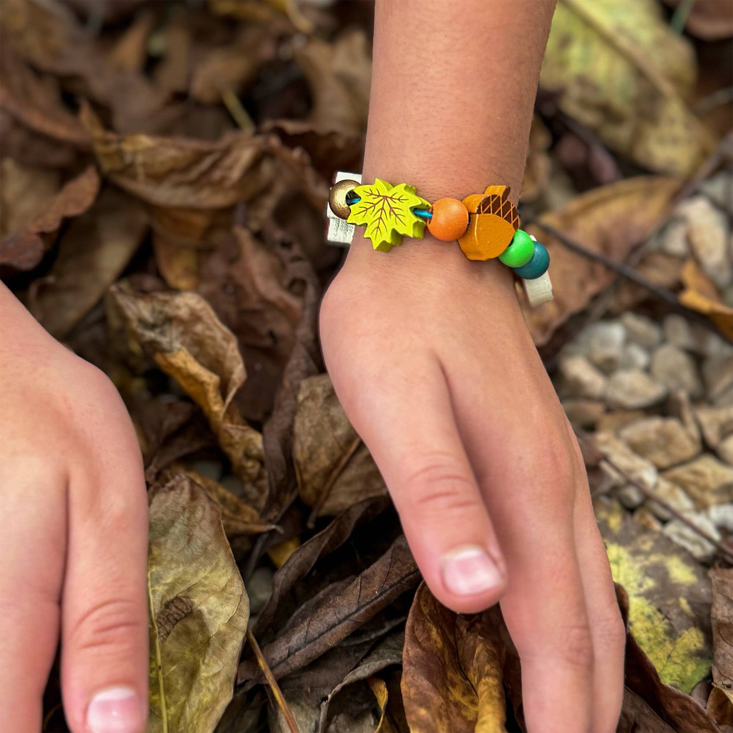Child's hand wearing a colorful beaded bracelet with a leaf charm on a background of dried leaves.