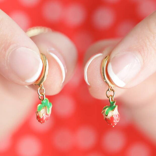 Gold hoop earrings with fruit-shaped charms held between fingers against a red background