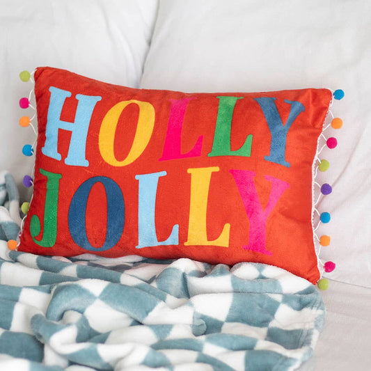 Colorful 'Holly Jolly' pillow on a patterned blanket with pom-poms.