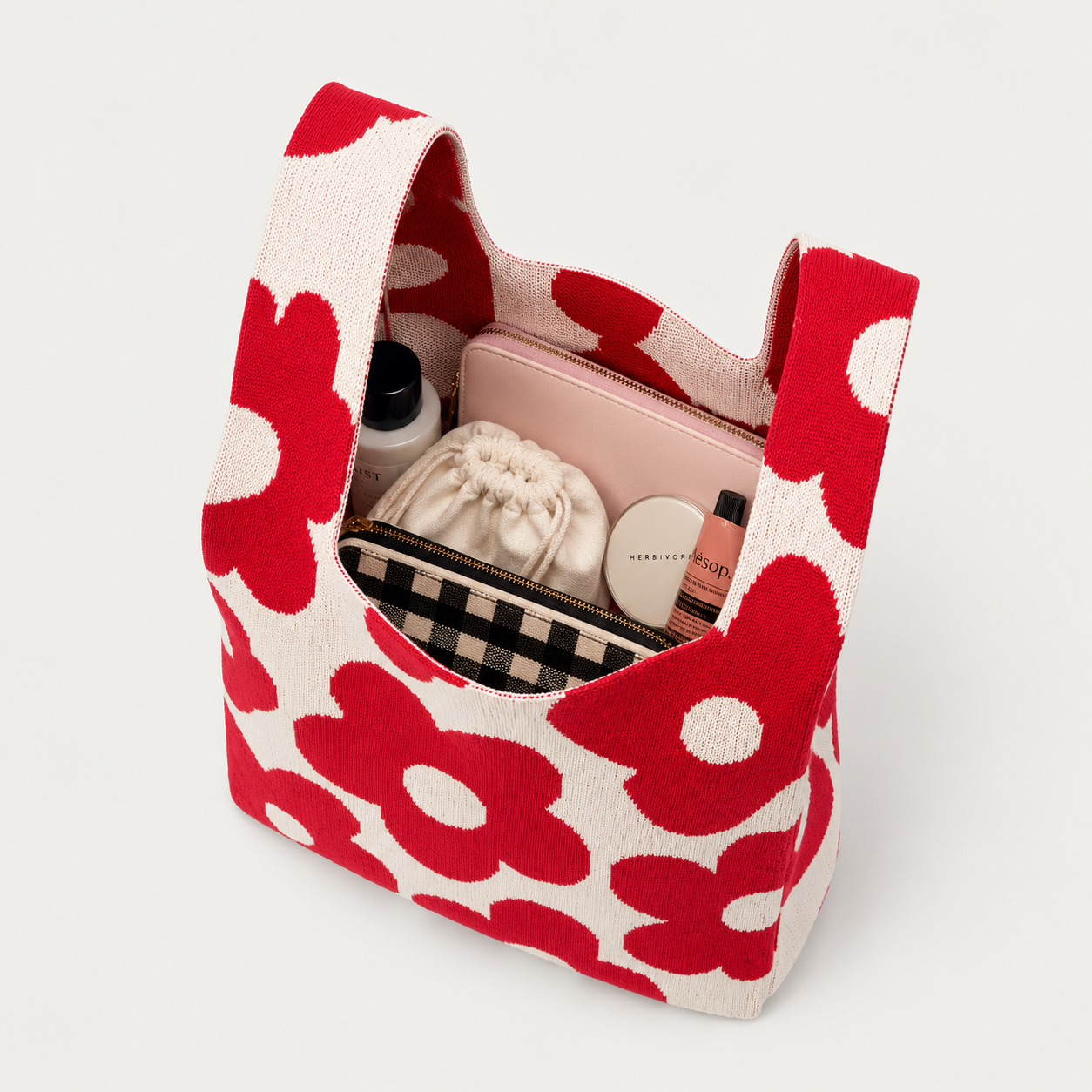 A tote bag with a red and off-white floral pattern, shown open and filled with various cosmetic items against a light gray background.