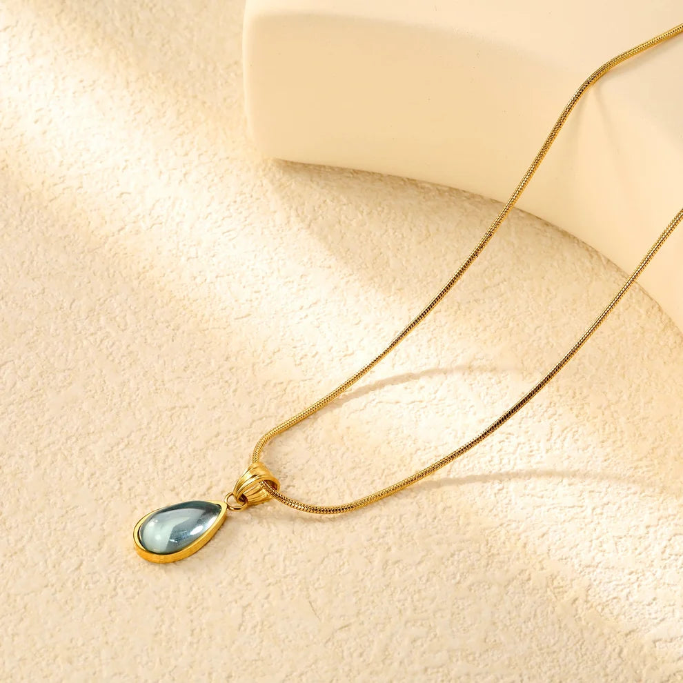 A gold chain necklace with a teardrop-shaped pendant featuring a light blue-gray stone in a gold bezel setting, displayed on a textured beige surface with soft shadows.