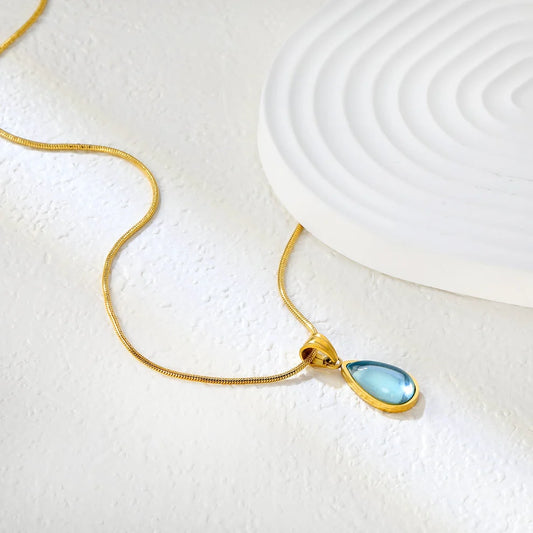 Gold necklace with a blue teardrop pendant on a textured white surface
