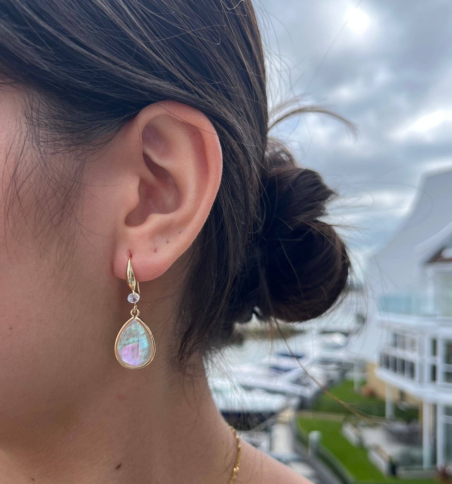 Close-up of an ear wearing a gold earring with a colorful gemstone, blurred outdoor background