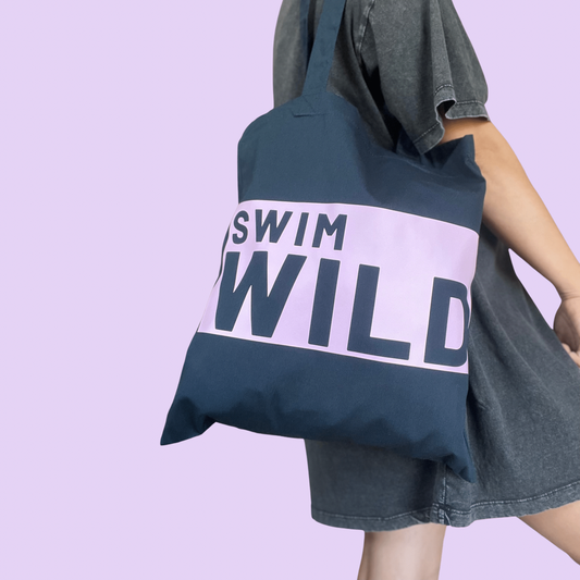 Person holding a tote bag with 'Swim Wild' text against a light purple background