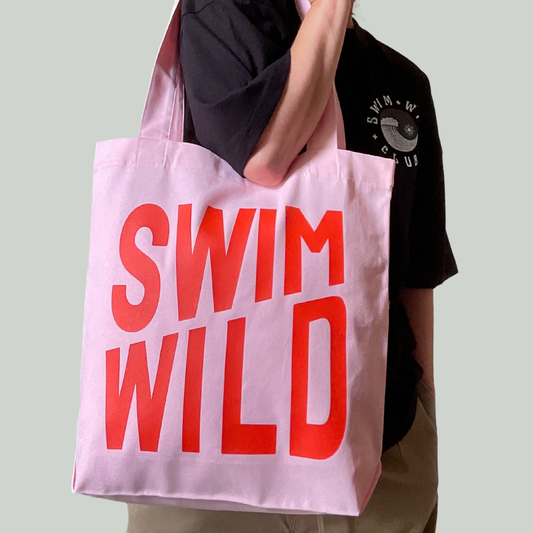 Person holding a pink tote bag with 'SWIM WILD' text against a plain background