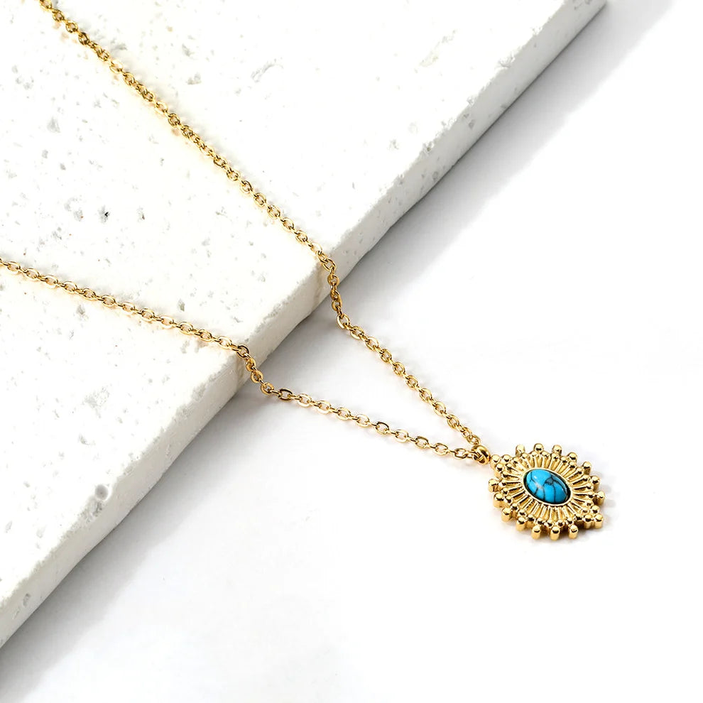 A delicate gold chain necklace with an intricate sunburst pendant featuring a central turquoise blue oval stone, displayed on a white textured stone slab against a white background.