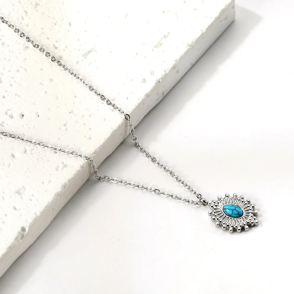 A silver-toned chain necklace with an evil eye pendant featuring a turquoise-blue center stone and silver radiating details, displayed on a white textured stone block against a white background.