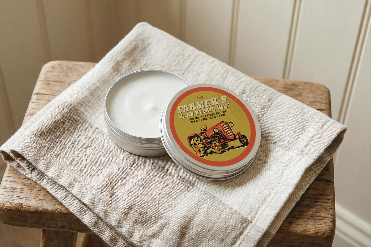 A round metal tin of hand repair wax sits open on a folded cream linen towel, placed on a rustic wooden stool against a light paneled wall.